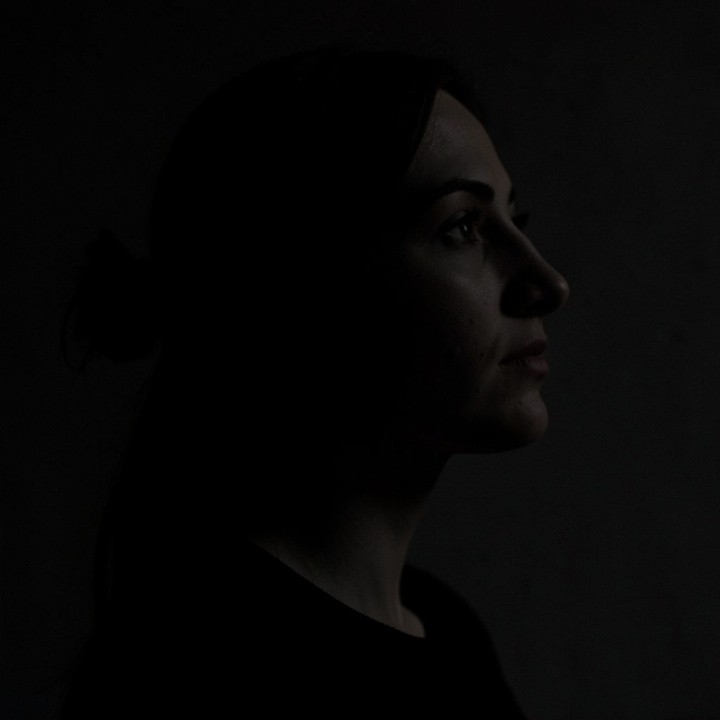 A warm, dark-toned portrait of a woman with a gentle smile, soft natural light falling across her face against a deep shadowed background.